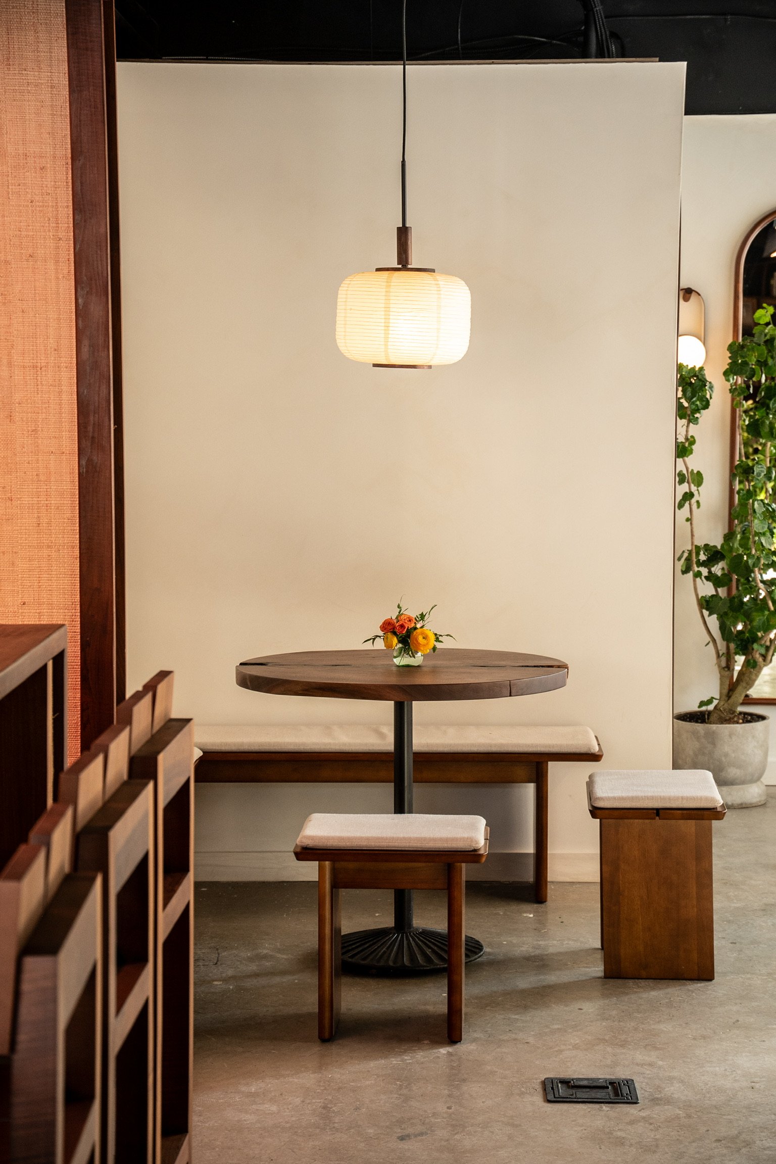 A cozy interior corner of a restaurant with a wooden table and seating, a hanging paper lantern light, a small vase with colorful flowers, a large potted plant, and a decorative mirror on the wall.