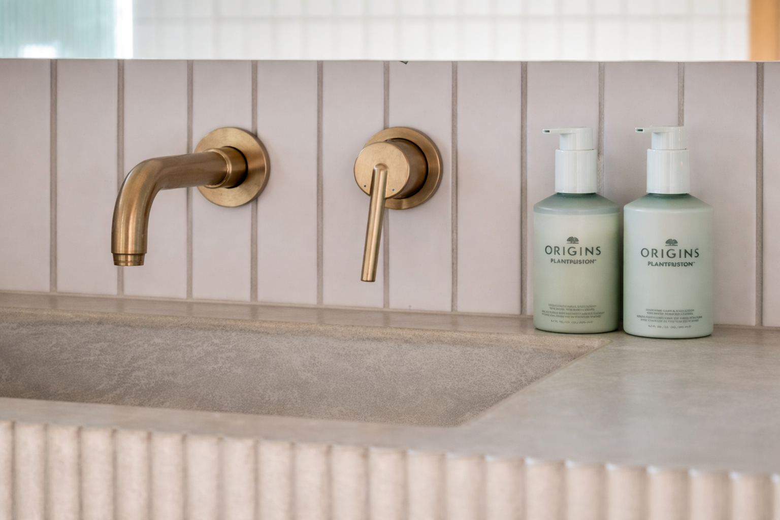 Close-up of a bathroom sink with a brass faucet and handle, two bottles of Origins skin care products, and white tiled wall.