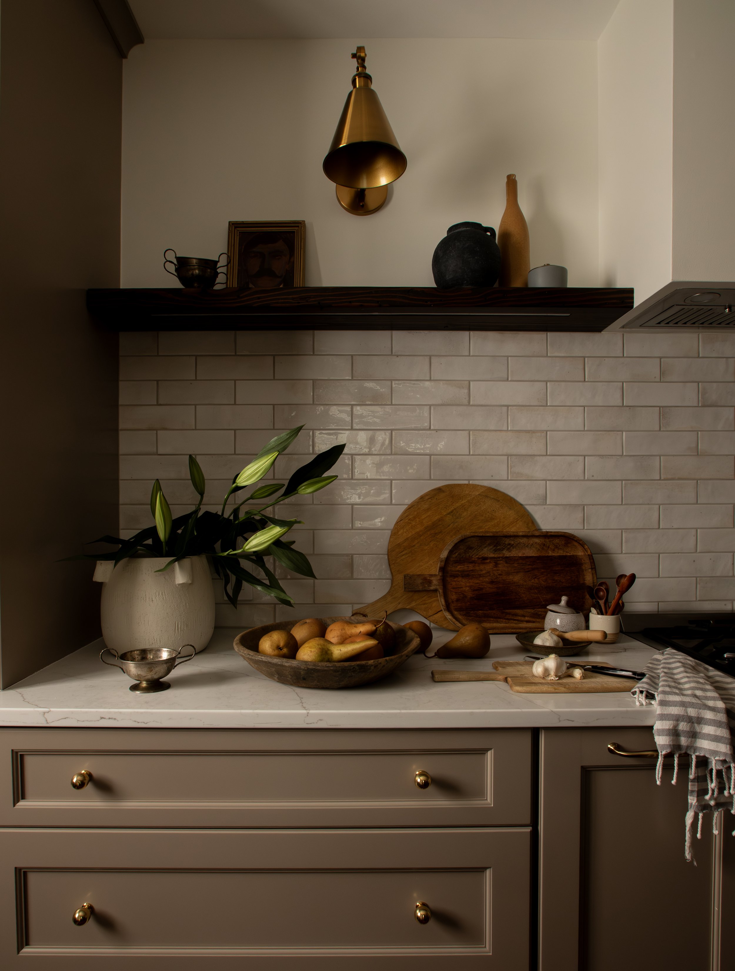 A kitchen countertop with a plant in a white vase, a bowl of potatoes, wooden cutting boards, garlic, and kitchen utensils, against a white brick backsplash and beige cabinets.