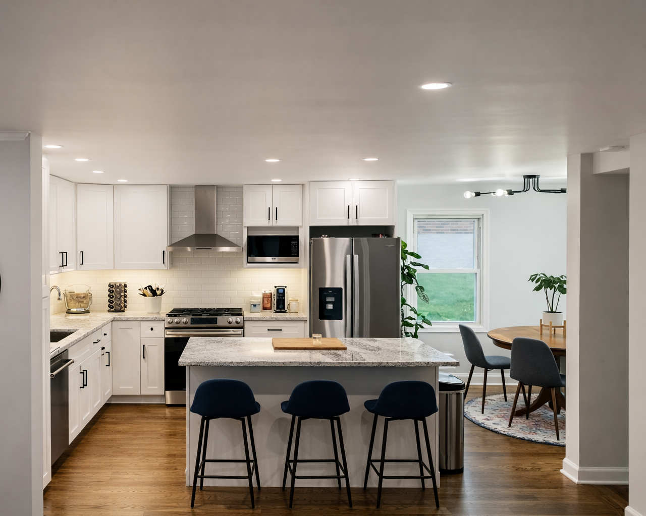 Modern white kitchen with granite island, stainless steel appliances, black barstools, and a dining area with a round table and chairs near a window with plants.