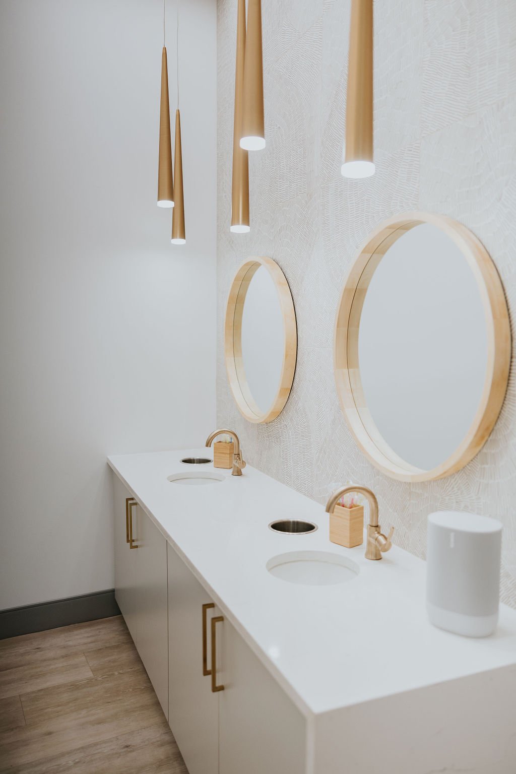 A modern bathroom with white vanity, three round mirrors with wooden frames, and hanging pendant lights with wooden shades.
