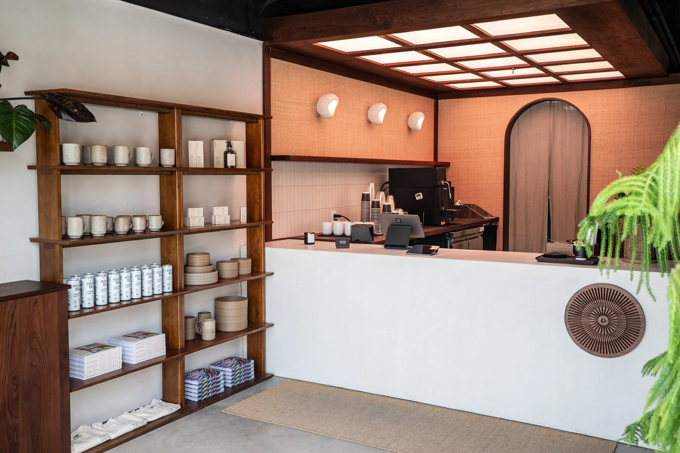 Interior of a cafe or store with a wooden counter, shelves with ceramic cups, mugs, plates, and magazines, a coffee machine, and a plant. The ceiling has a grid pattern with lighting, and there's a large wooden door with an arched top in the backgrou