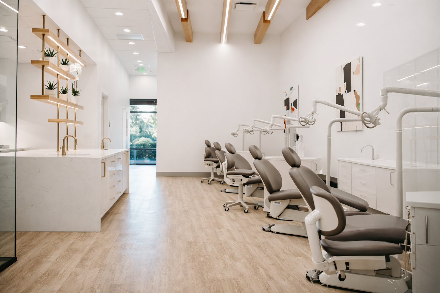 Modern dental treatment room with dental chairs, white cabinetry, artwork on the wall, and a large window with a view of greenery outside.