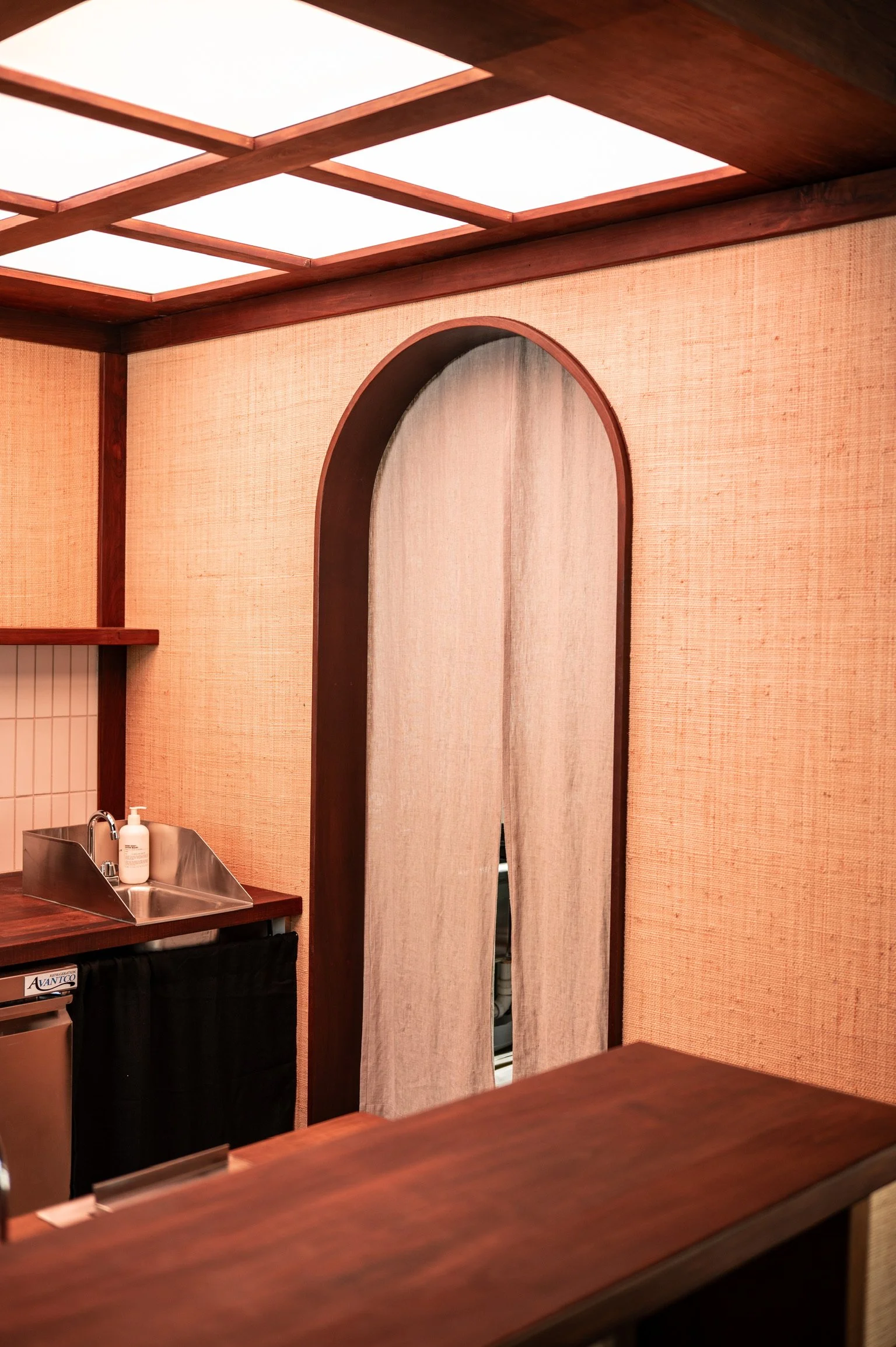 Interior of a room with wooden walls and ceiling, a mirror with a wooden frame, a stainless steel sink with soap, and a wooden countertop.