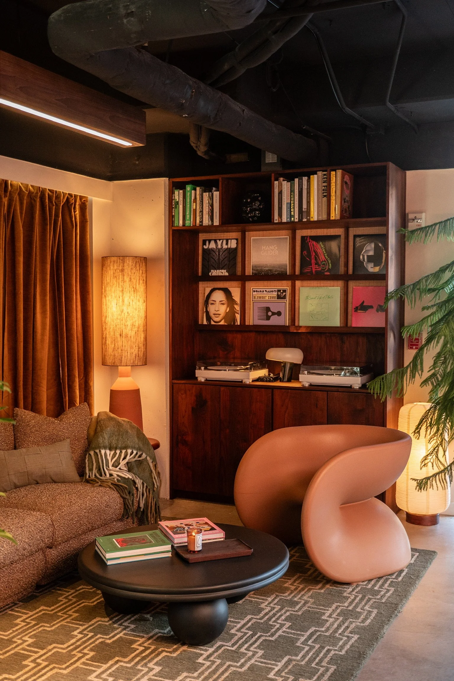 Living room with tan sofa, black round coffee table, pink modern chair, wooden bookshelf with books and records, amber curtains, warm lamps, green plant, and geometric patterned rug.