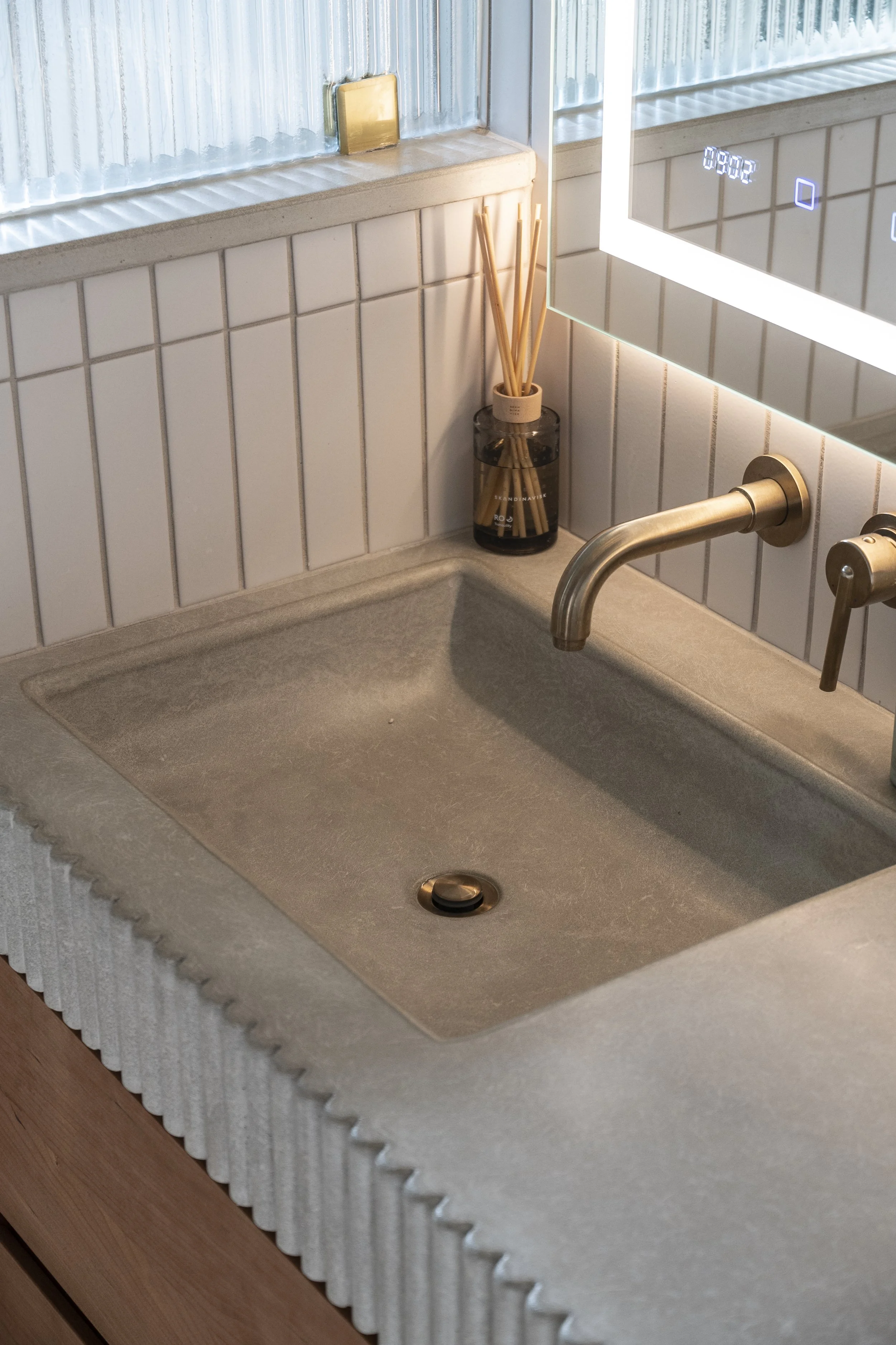 A kitchen sink with a beige countertop, a gold-tone faucet, and a glass jar with bamboo sticks, next to a mirrored cabinet with an LED light.