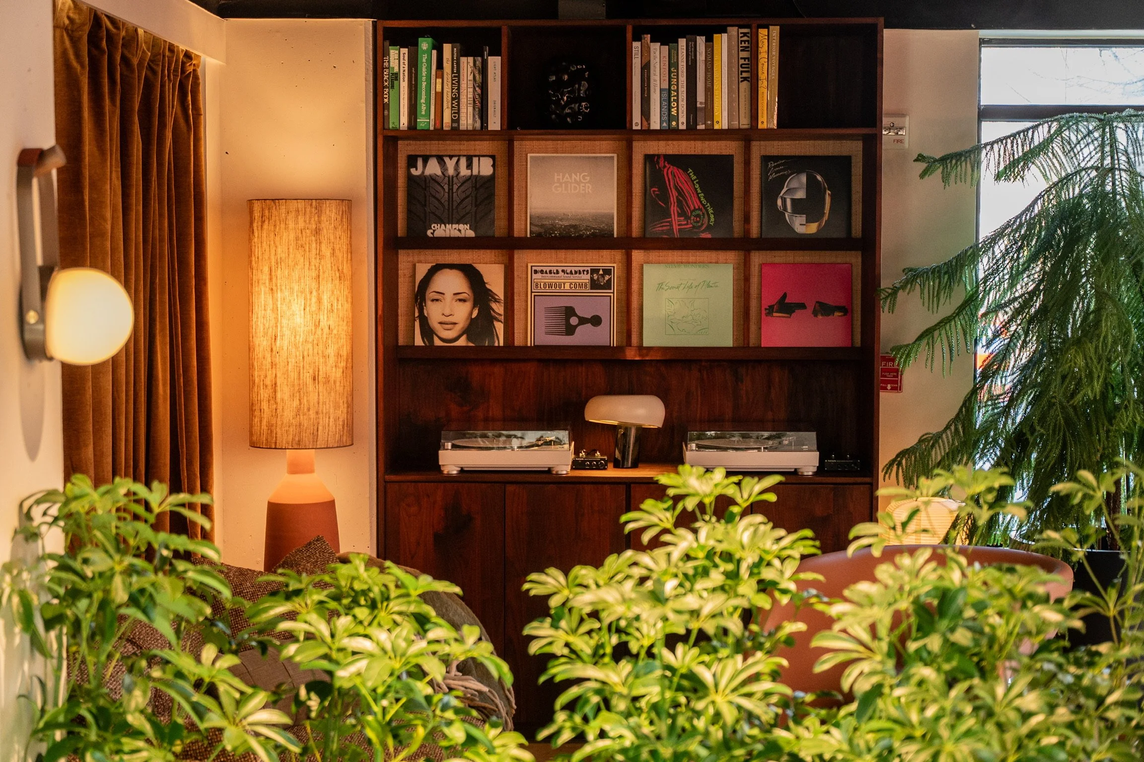 A cozy living room corner with a tall beige lampshade, a wooden bookshelf filled with books and vinyl records, green plants in the foreground, brown curtains, a wall-mounted light fixture, and a window with visible trees outside.