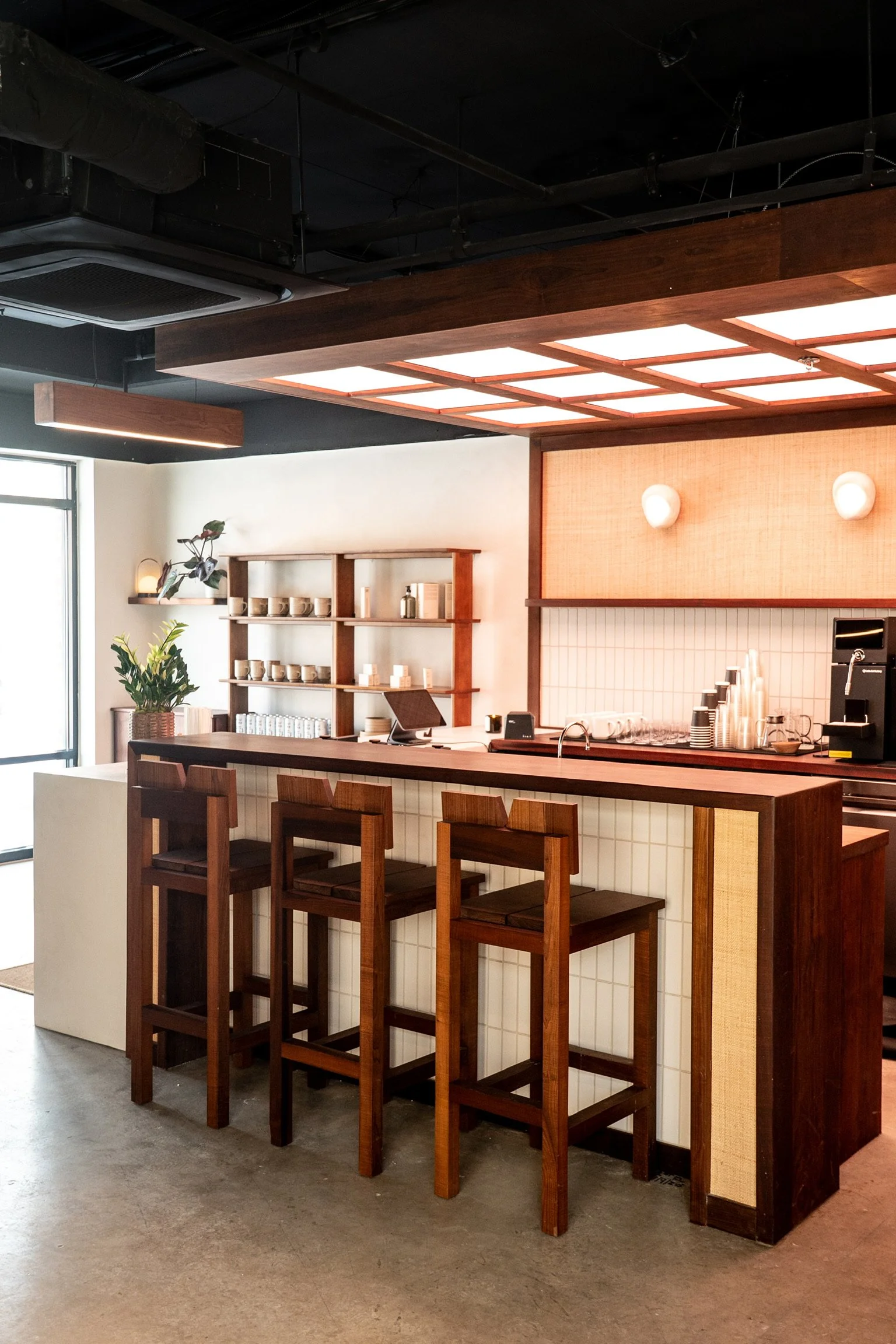Interior of a modern coffee shop featuring a bar counter with four wooden chairs, a shelf with cups and books, a small potted plant, and a coffee machine, with a ceiling decorated with wooden panels and soft wall lighting.