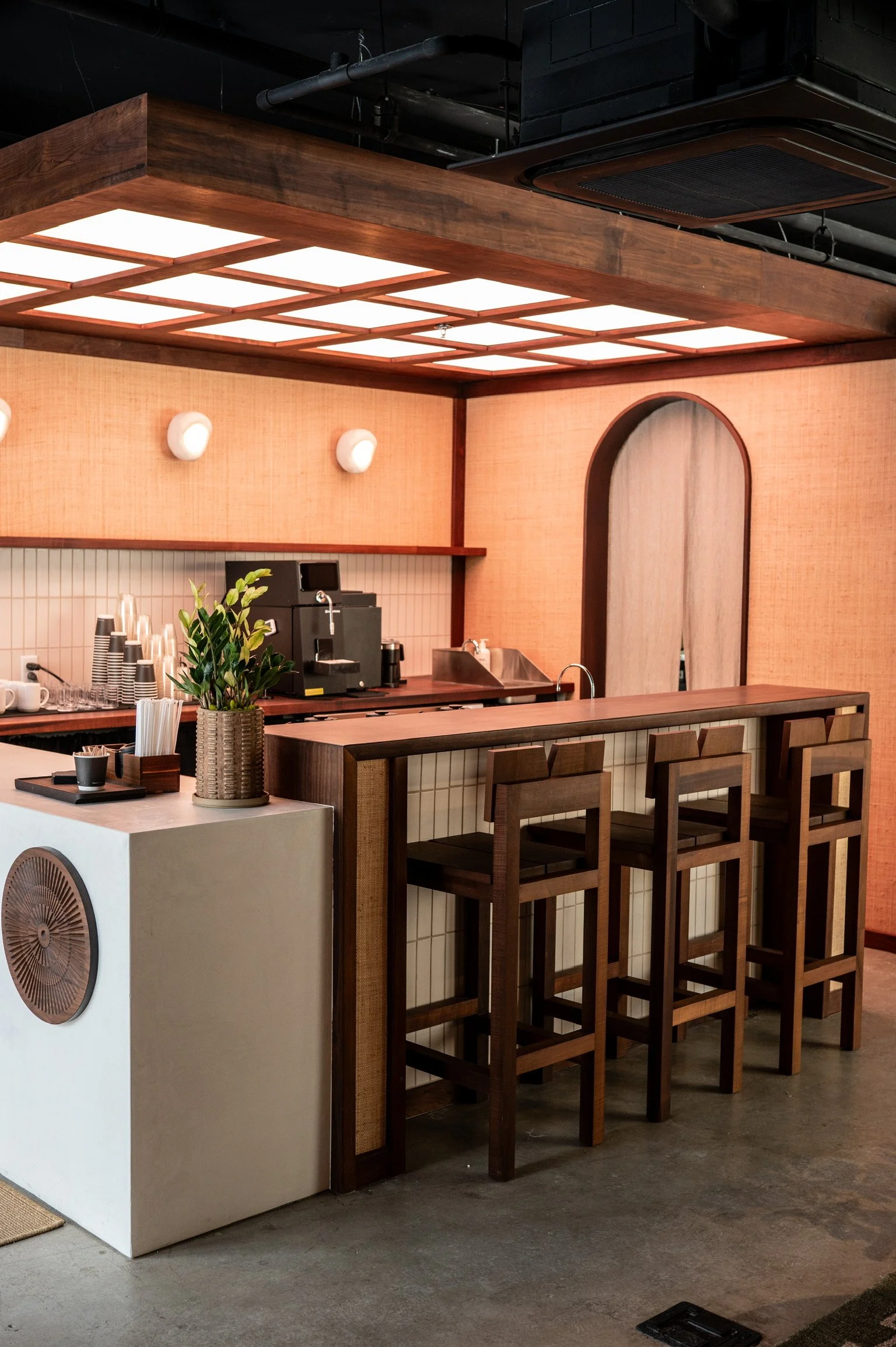 A coffee bar area with a white counter, potted plant, and bar stools, featuring a wooden ceiling structure and a barista station with a coffee machine.