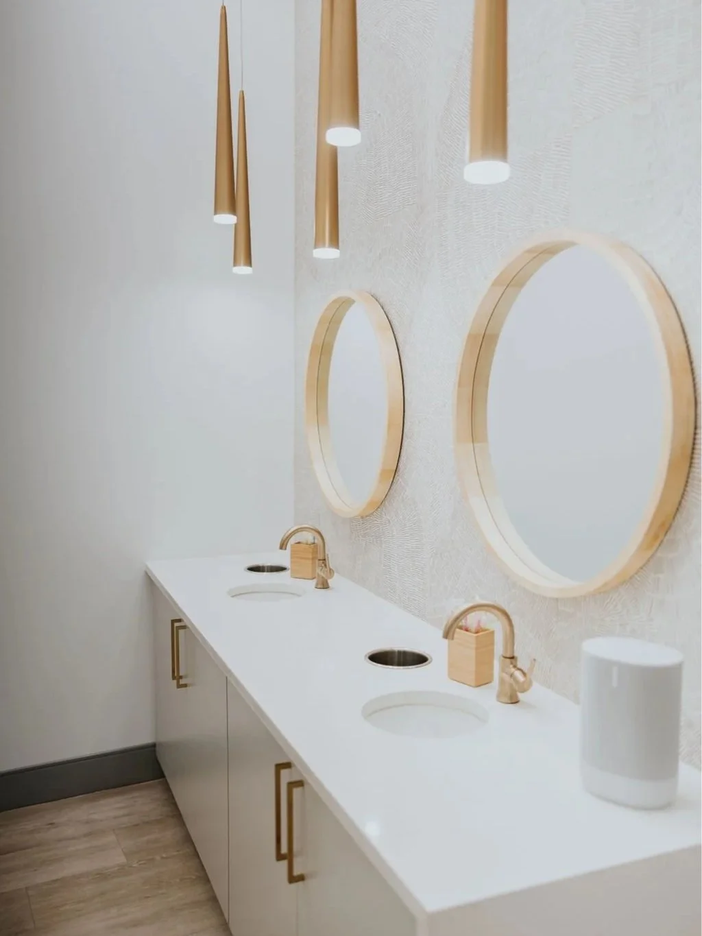 A modern bathroom with a white countertop, three round mirrors with wooden frames, two sinks, gold faucets, and three hanging pendant lights.