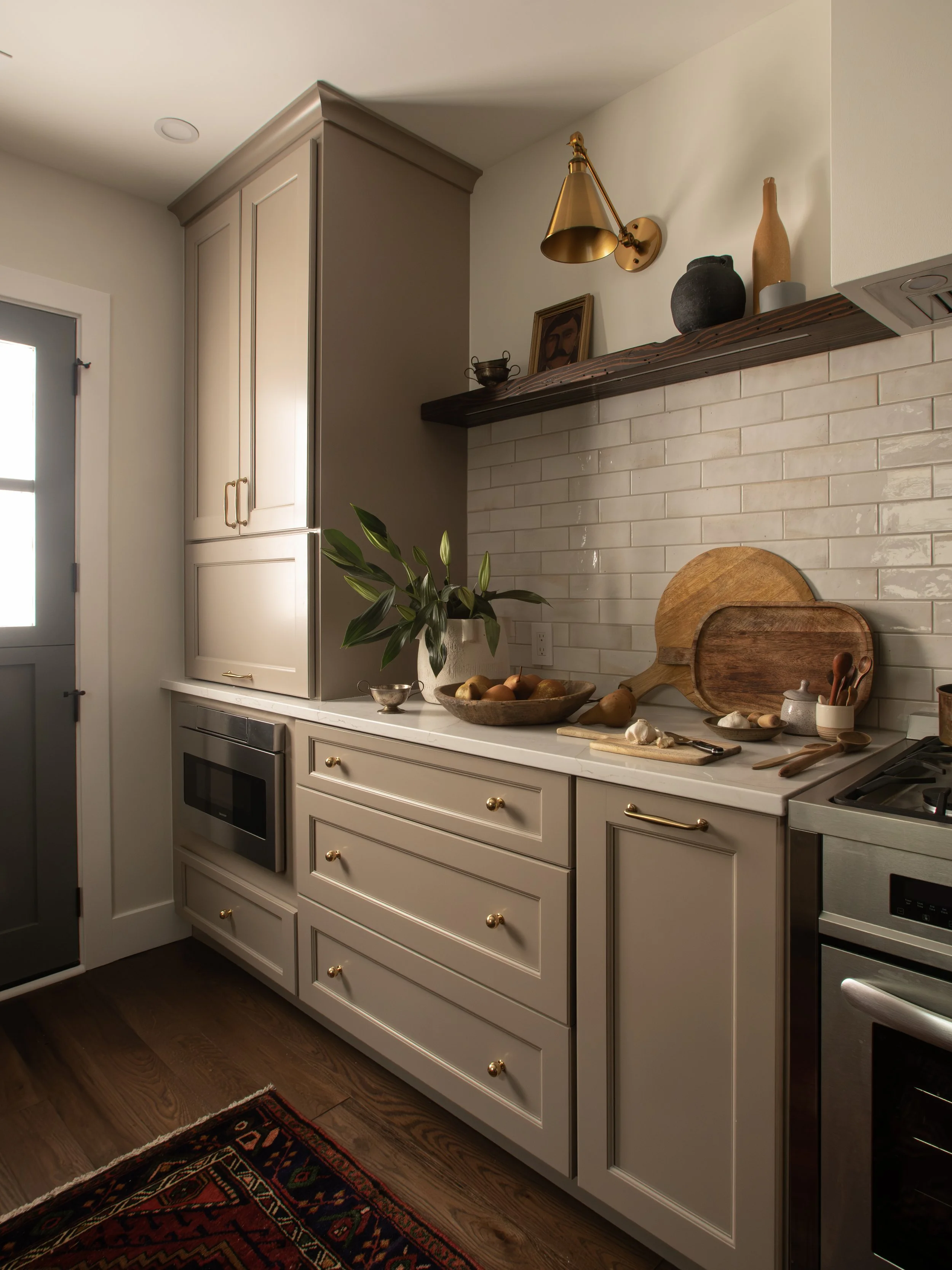 Kitchen countertop with chopping boards, garlic, and utensils, beige cabinets with gold knobs, plant in a white vase, brick backsplash, upper shelf with decorative items, gold wall sconce, closed door with window.