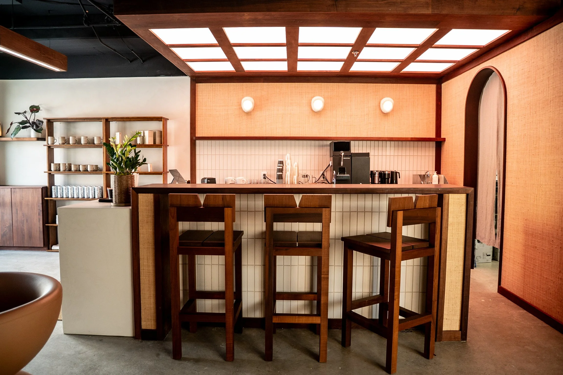 Interior of a cafe or restaurant with a bar counter, wooden stools, shelves holding cups and books, and decorative lighting, featuring a warm color palette of browns and beige.