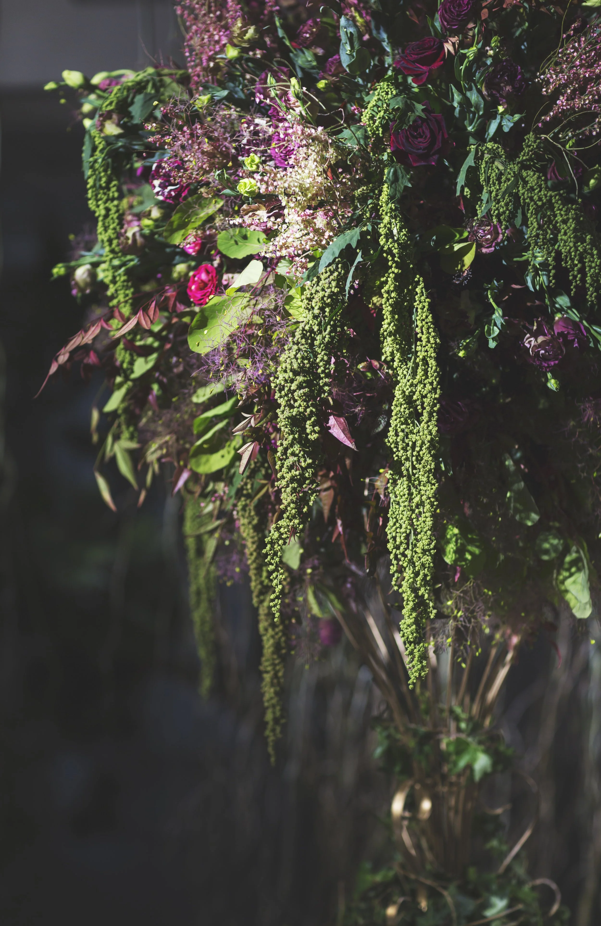 Lush floral arrangement with green foliage, pink and maroon flowers, and hanging greenery in a dark background.