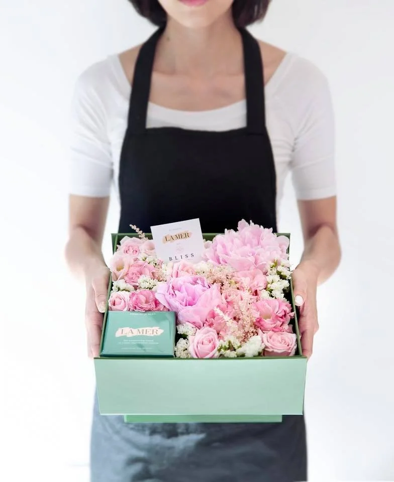 Person holding a green gift box filled with pink roses and flowers, containing La Mer skincare products.