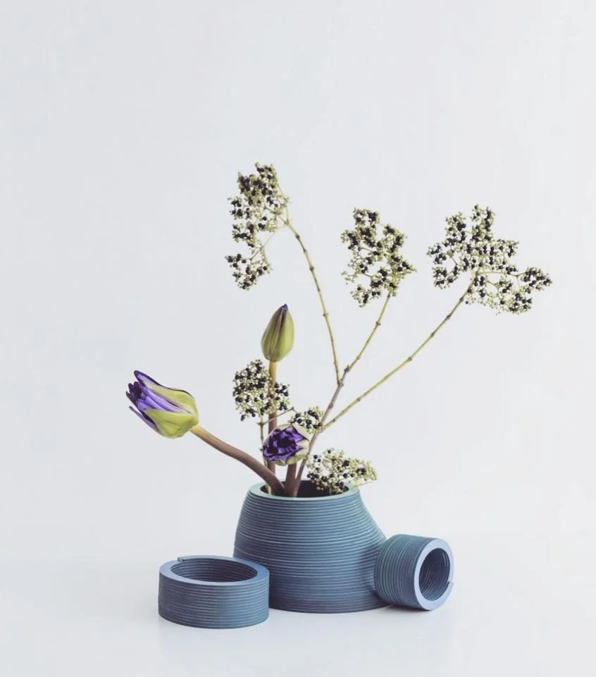 Purple and green flowers in a uniquely shaped blue vase with additional blue cylindrical pieces on a white background.