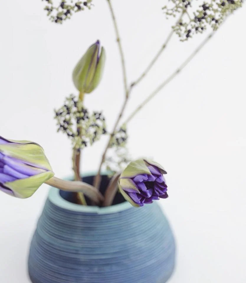 Close-up of purple tulip buds with green accents in a textured blue vase, surrounded by delicate branches and small white flowers against a white background.