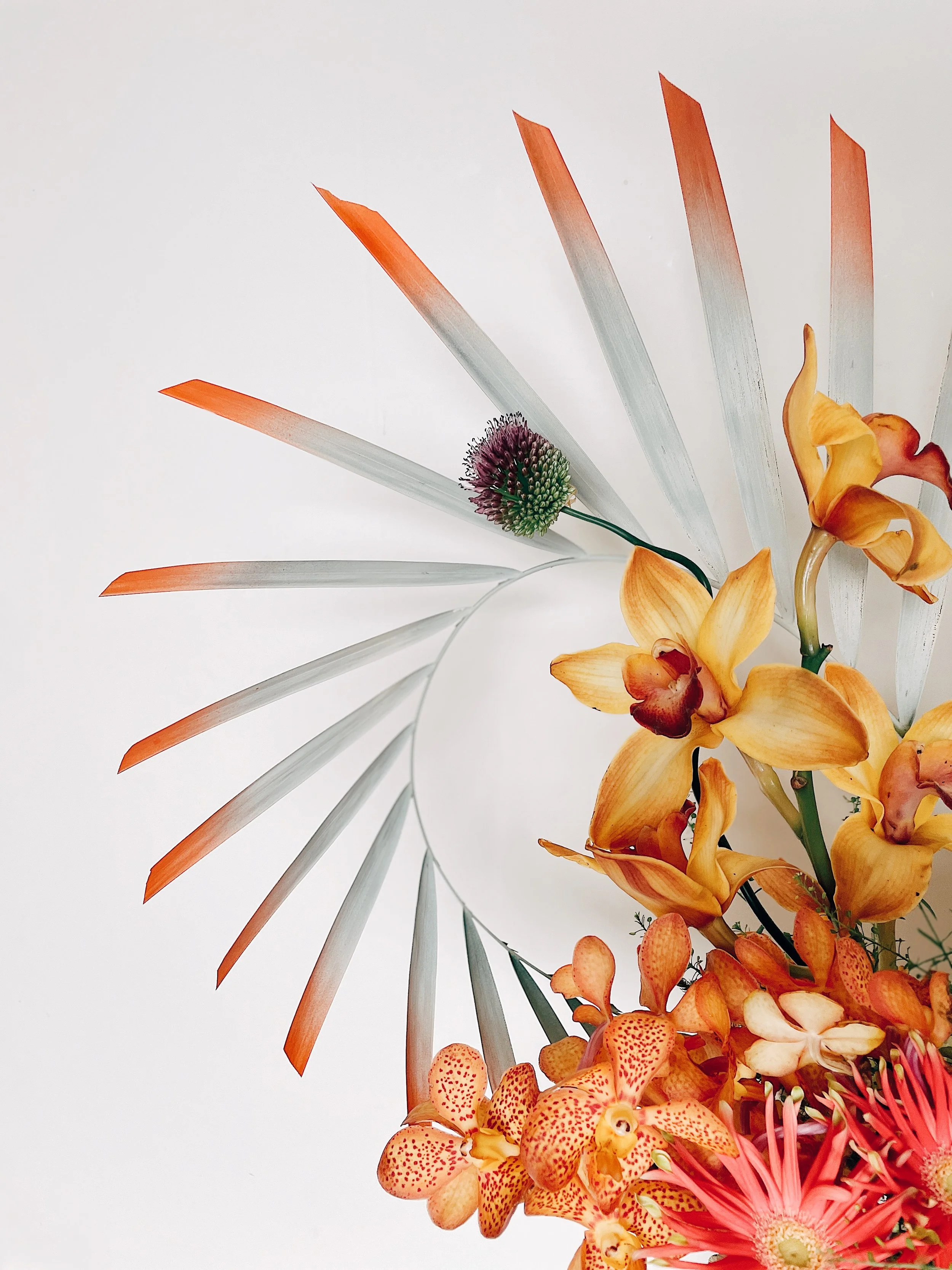 Abstract floral arrangement with orange and yellow lilies, spiky green and pink flower, and orange ombre leaves fanned around a circular mirror on a white background.