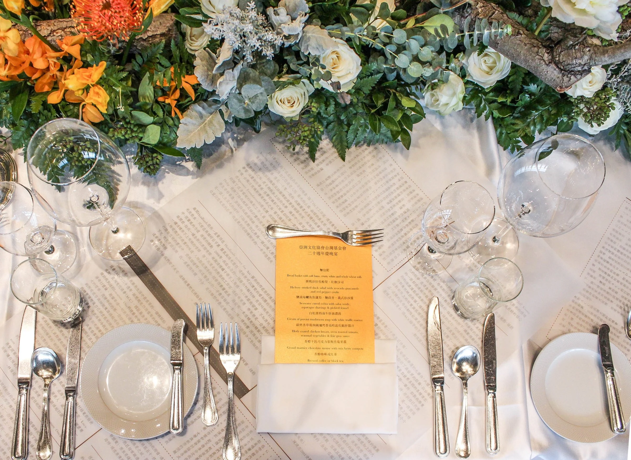 Elegant table setting with silverware, wine glasses, and a menu card on a white tablecloth, decorated with a floral arrangement of green leaves, white roses, orange flowers, and a tree branch.