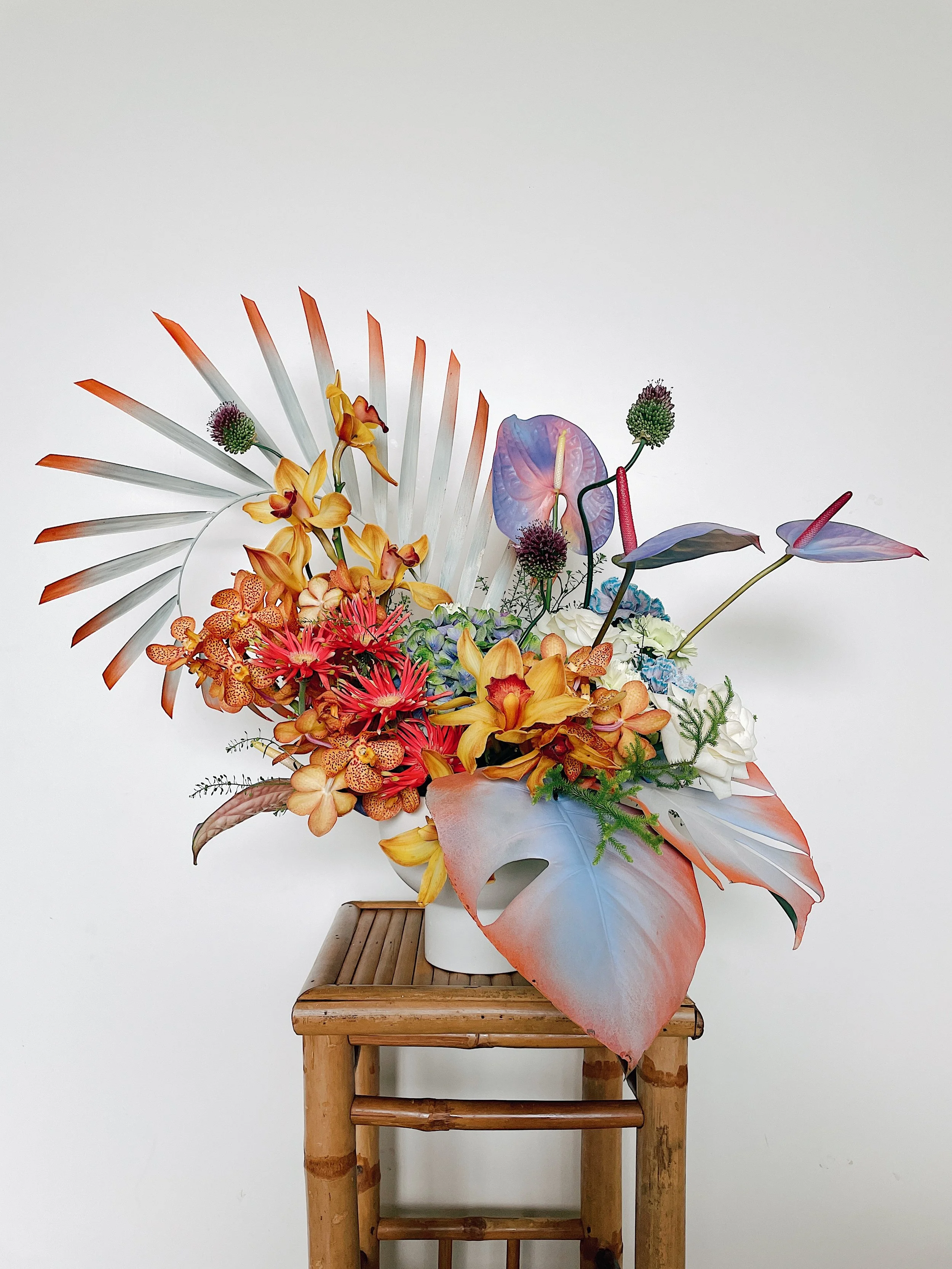 Artistic floral arrangement with tropical and exotic flowers on a bamboo stool, featuring colorful leaves and blossoms against a white background.