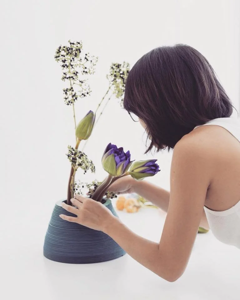 Person arranging purple flowers and white blossoms in a blue vase.