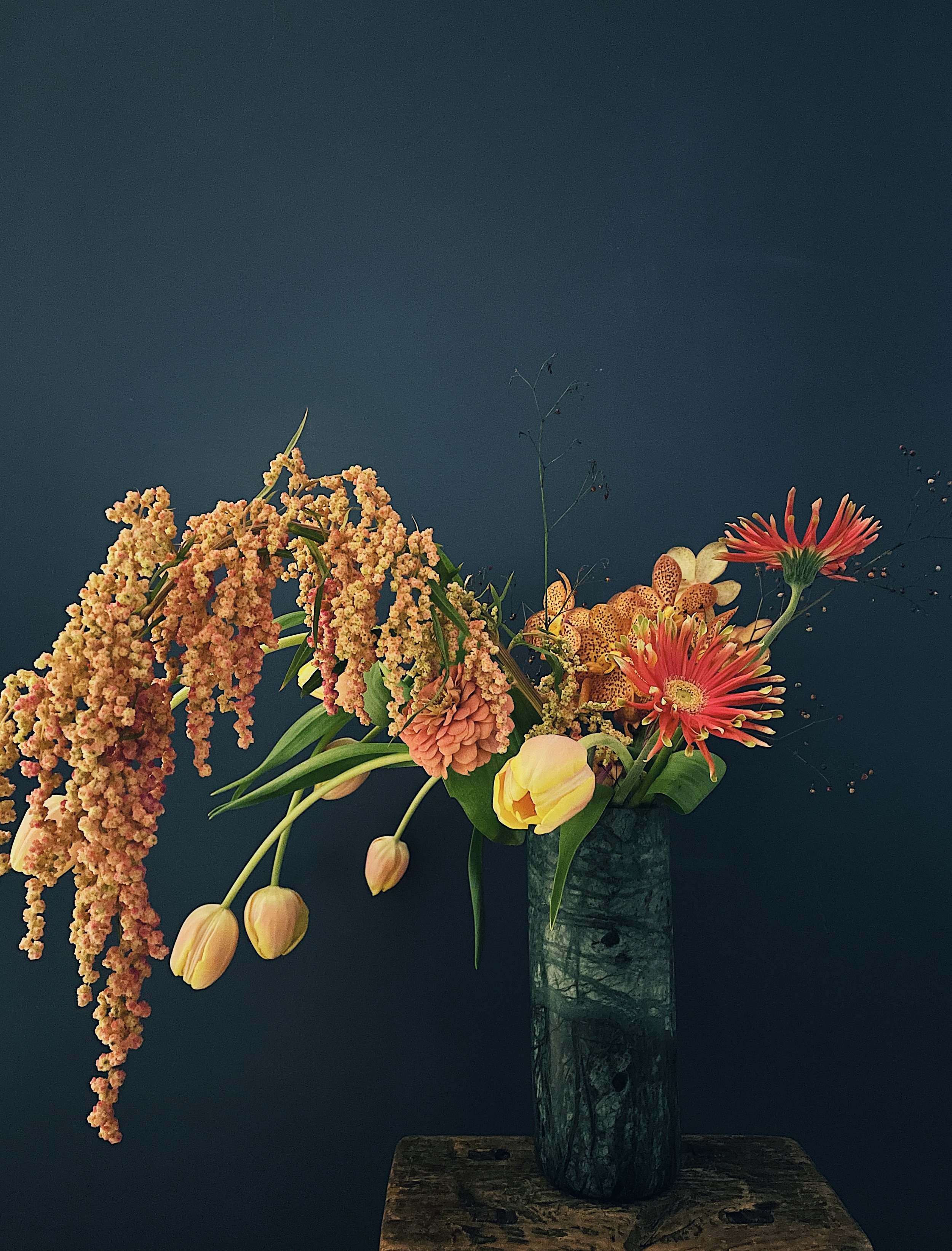 Elegant floral arrangement in a dark vase against a dark background, featuring tulips, gerbera daisies, and cascading blooms.