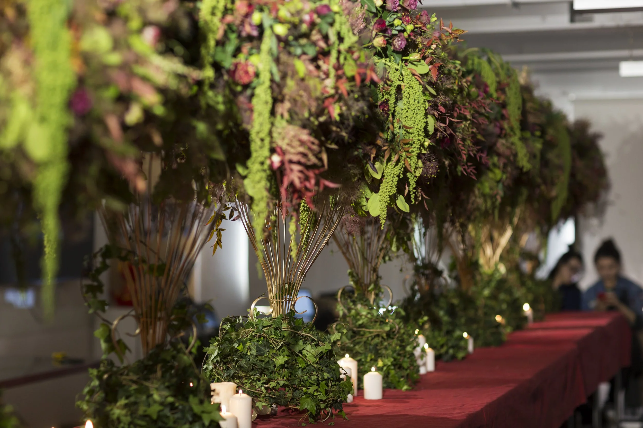 Elaborate floral arrangements with greenery on tall stems above a red tablecloth, surrounded by lit white candles, creating an elegant and intimate setting.