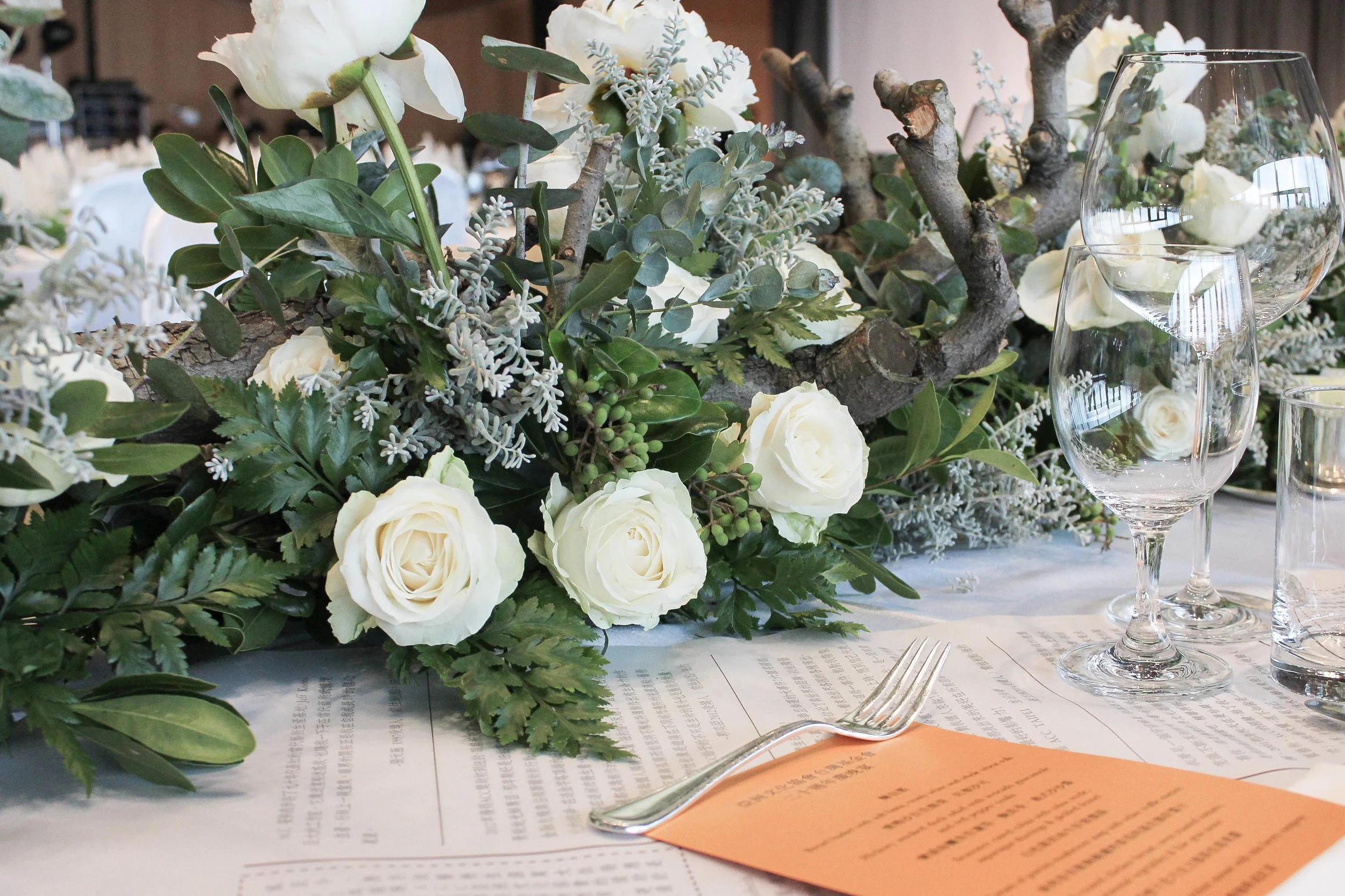 Table setting with a centerpiece featuring white roses, greenery, and branches, accompanied by wine glasses and an orange menu.