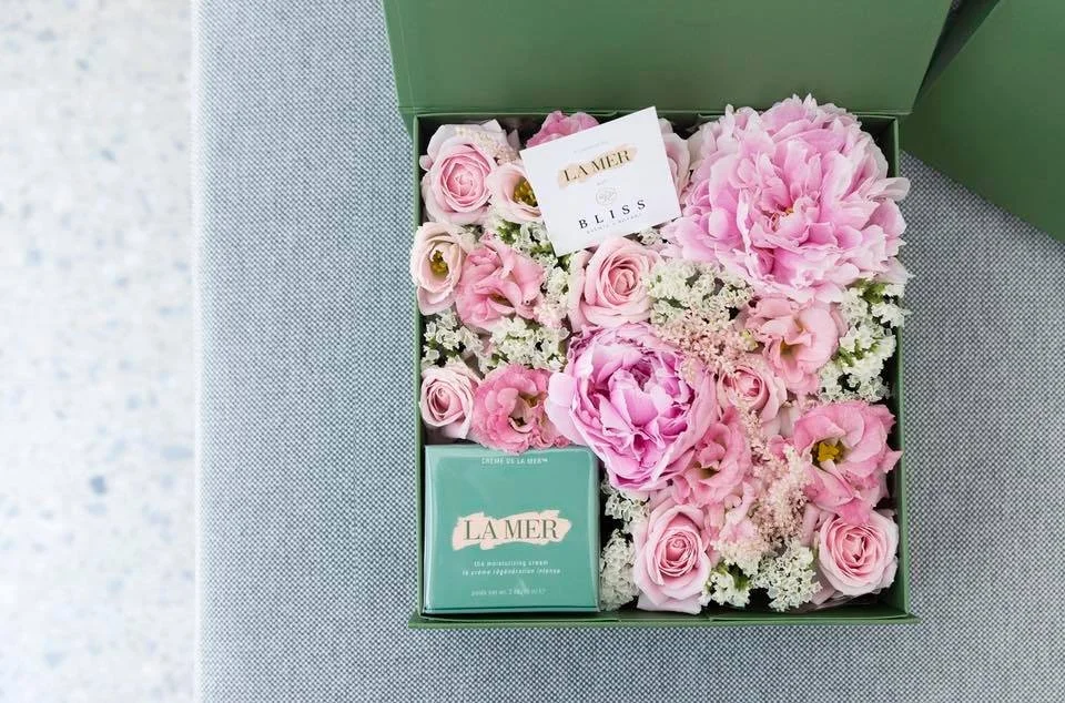 Gift box with pink flowers, La Mer skincare product, and a card on gray surface.