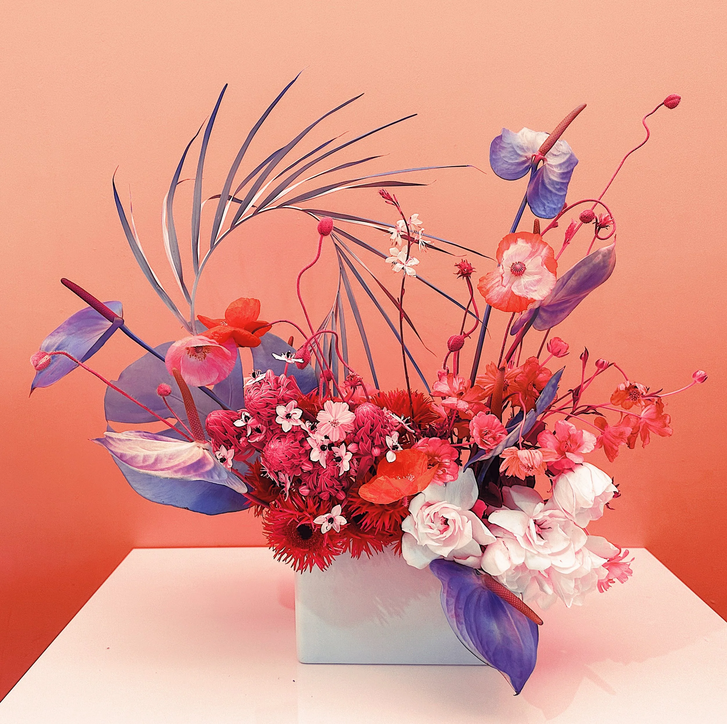 Vibrant floral arrangement with pink, red, and purple flowers and leafy accents in a rectangular vase against a peach-colored background.