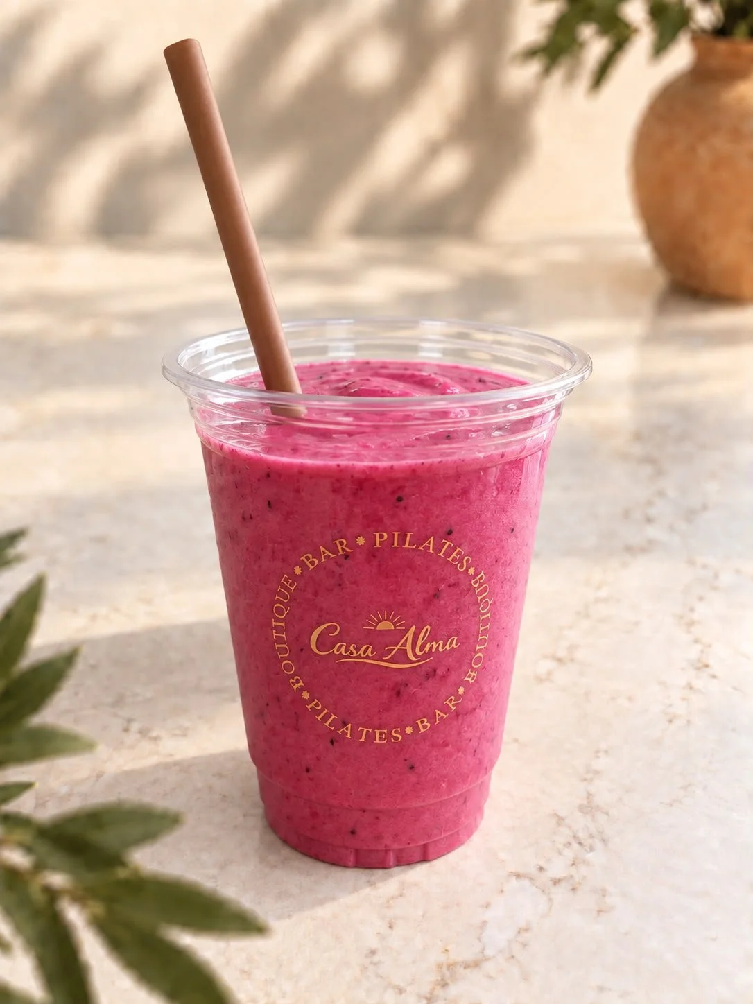 Pink Alma Colada Smoothie; Pink Pitaya, Mango, Kokos, Banaan, Dadel in een doorzichtige plastic beker met Casa Alma erop, en een rietje erin