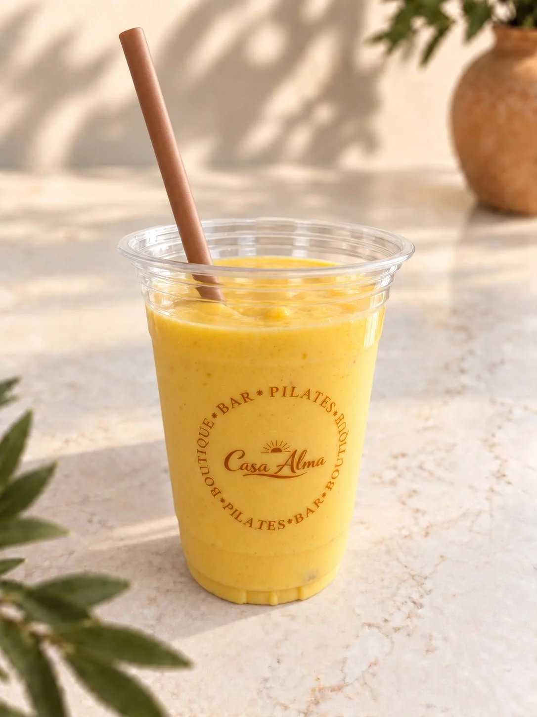 Gele Alma Sunshine Smoothie; Kokos, Acerola, Dadel, Passievrucht, Ananas, Mango in een doorzichtig bekertje met een roerstaafje, op een lichte, natuurlijke achtergrond met een vaas en groene bladeren op de achtergrond.
