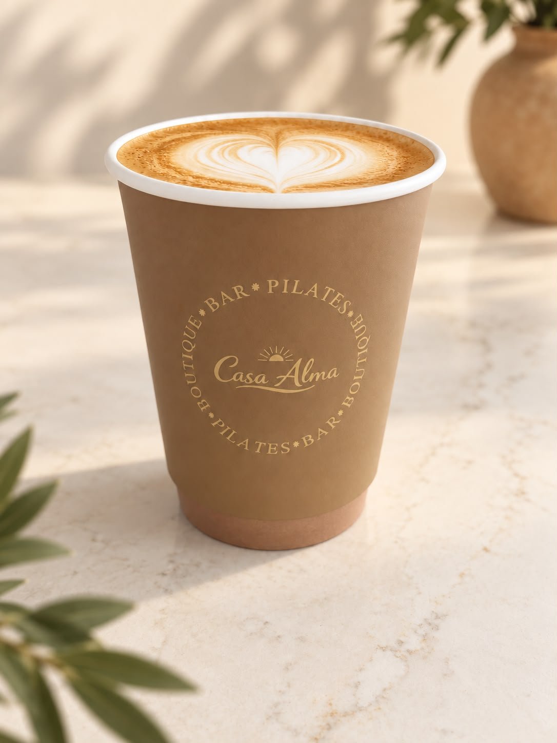 Een papieren bekertje met cappuccino en latte art, met de tekst 'Casa Alma' en omliggende woorden zoals 'BOUTIQUE', 'BAR', 'PILATES' op een lichte, marmeren tafel.