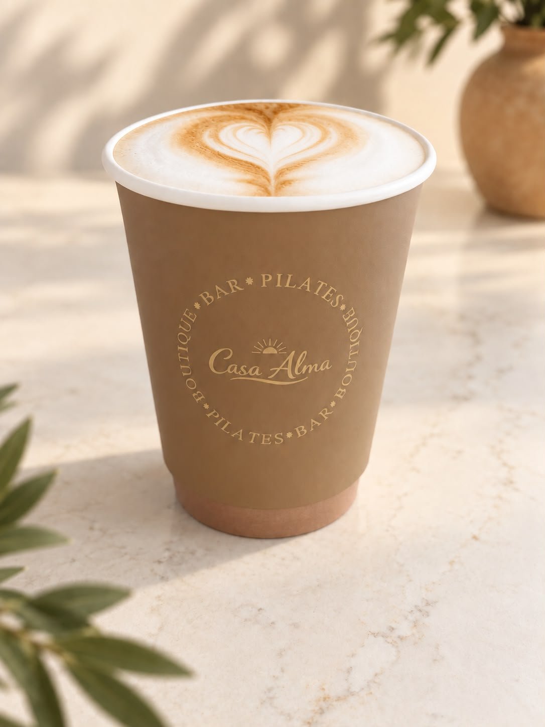 Een grote kop latte macchiato met schuim en hartvormige latte art op een beige papieren bekertje met tekst 'Casa Alma' en 'BOUTIQUE BAR PILATES'.