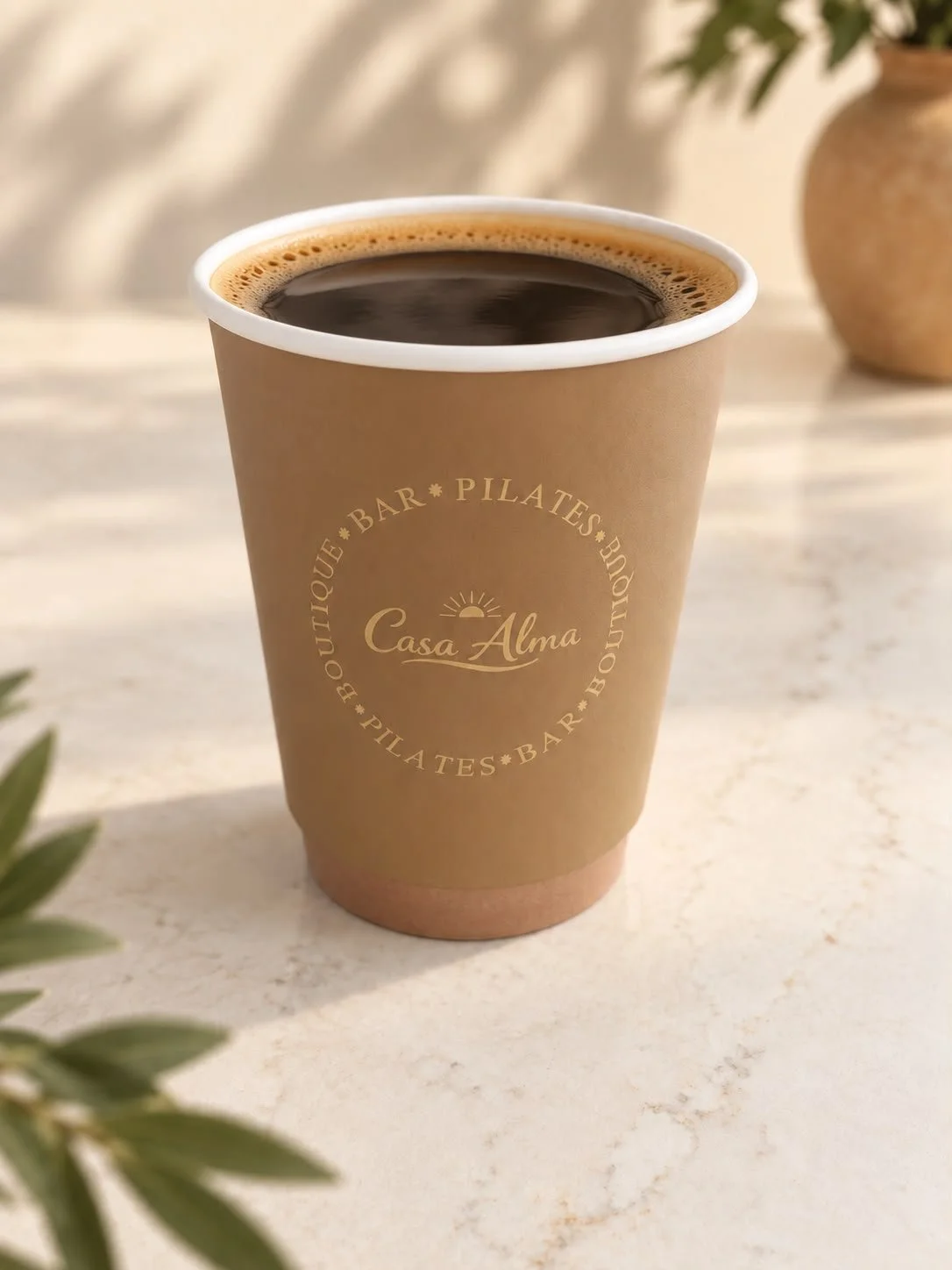 Een kop koffie in een papieren bekertje met het logo van Casa Alma, gelegen op een lichte tafel, met een vaas en groene bladeren op de achtergrond.
