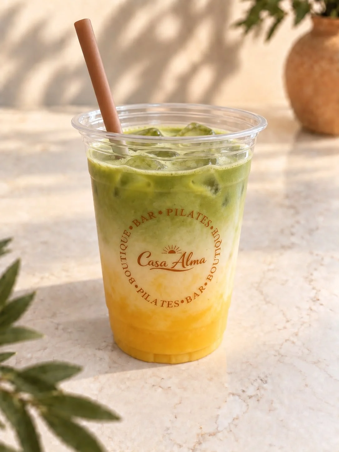 Klare plastic beker met een fijne gele en groene kleur en , gevuld met Iced Matcha Mango Latte en een roze rietje, op een lichte tafel met een vaas met planten op de achtergrond.