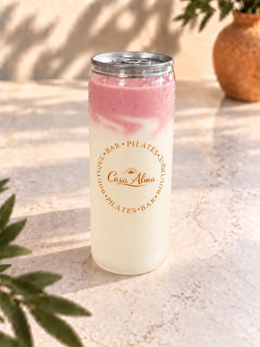 Een drinkfles met een logo van Casa Alma, gevuld met een twee-kleurige drank; Alma's proteïne power shake met pink matcha, op een lichte, zonnige tafel met een keramische pot en groene bladeren op de achtergrond.