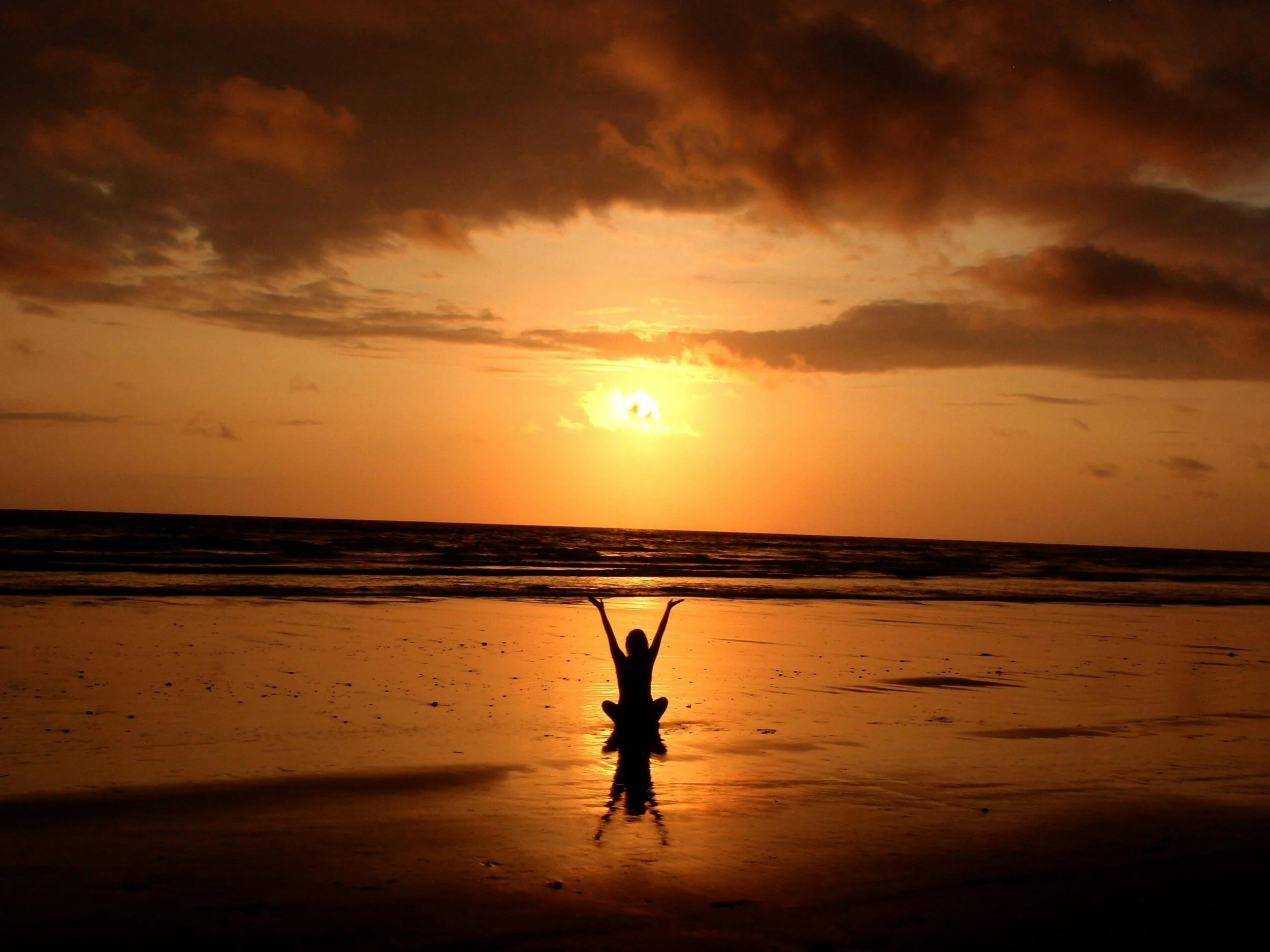 Silhouette of a person practicing yoga on the beach at sunset with arms raised, ocean waves in the background, and a vibrant orange sky with clouds.