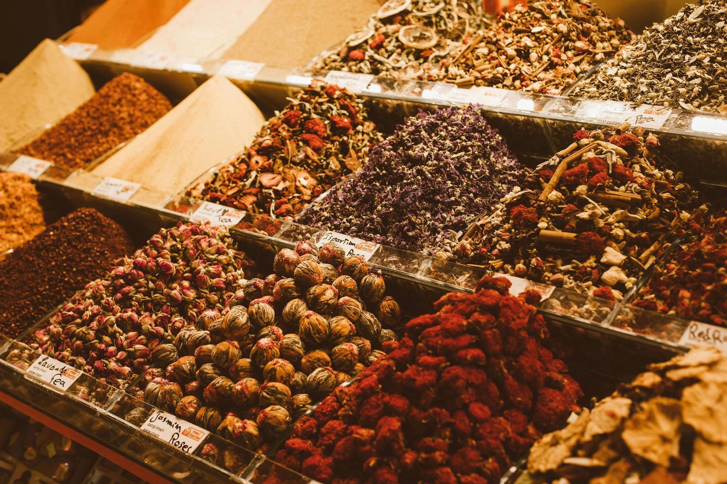 Display of various dried herbs, flowers, and spices in a market stall, arranged in separate bins with labels.