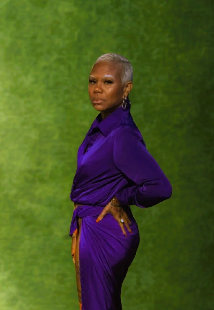 A woman with short gray hair, wearing a purple outfit with a matching skirt, is standing with one hand on her hip against a green background.
