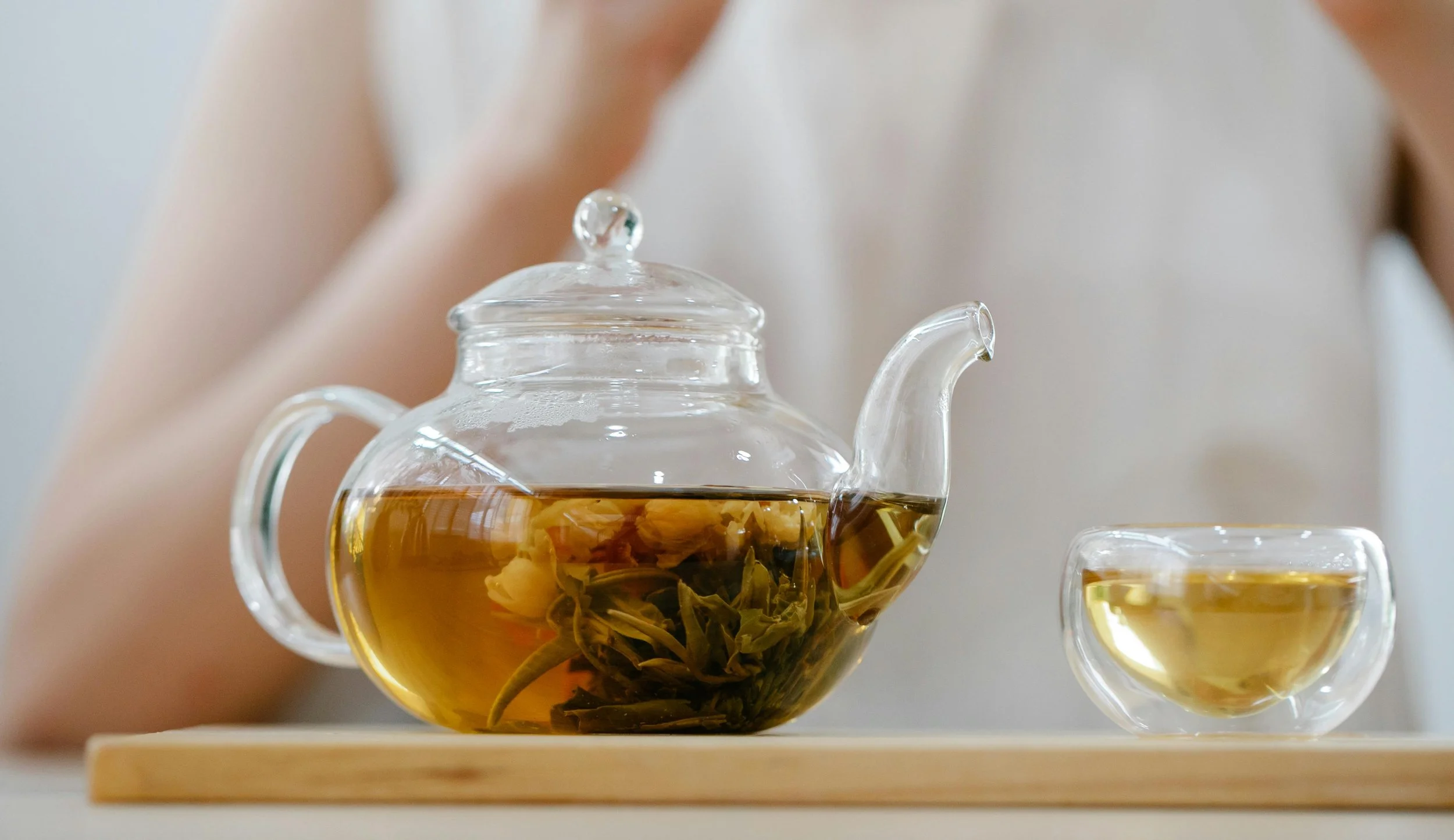 Glass teapot with loose leaf tea and a small glass teacup filled with tea on a wooden tray.