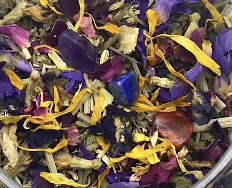 Close-up of a mixture of dried flower petals, leaves, and small stones or crystals in vibrant purple, yellow, pink, and blue colors.
