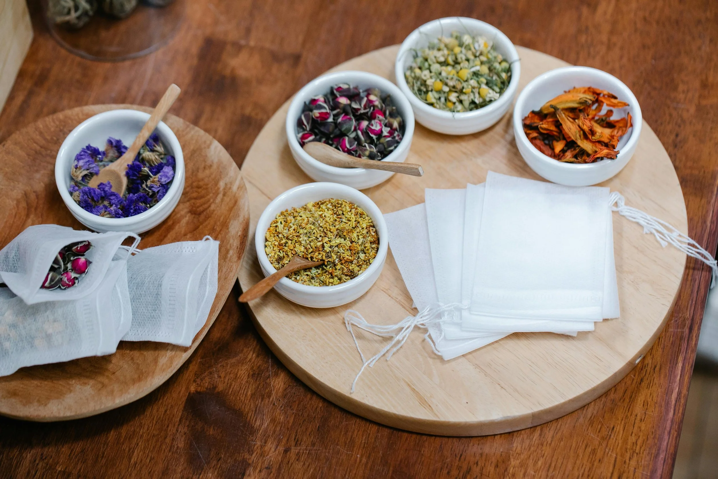 Assorted dried herbs and flowers in small white bowls on wooden trays, with white face masks nearby.