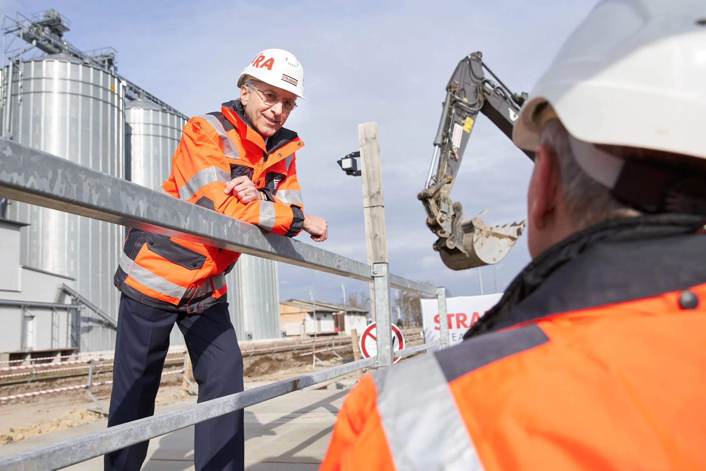 Zwei Bauarbeiter in Sicherheitskleidung bei einer Baustelle mit Silos und Baugeräten.