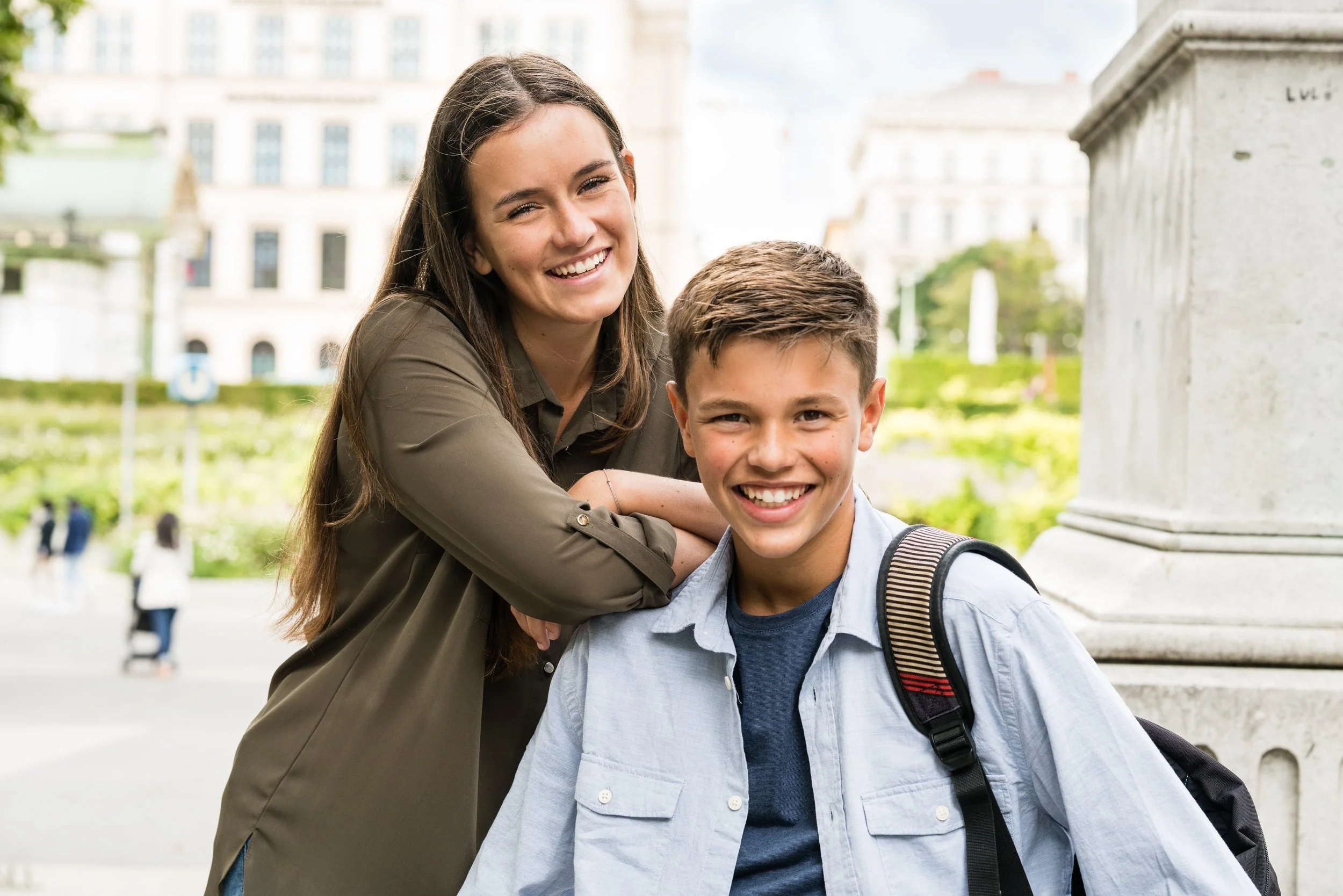 Ein lachendes junges Mädchen und ein lachender Junge mit Rucksack im Freien vor einem Gebäude.