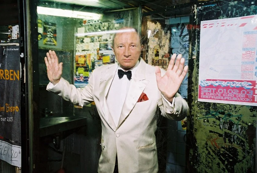 Ein Mann in einem weißen Anzug mit Fliege und roter Einstecktuch steht hinter einer Glasfassade, hebt die Hände in die Luft, in einem graffitiüberzogenen Raum mit Posters und Graffiti an den Wänden.