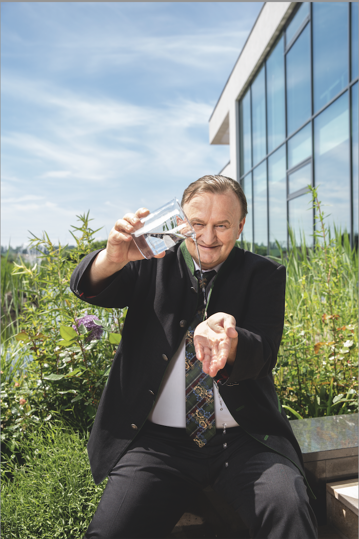 Ein Mann in einem schwarzen Anzug mit Krawatte sitzt im Freien vor einem modernen Glasgebäude und gießt Wasser aus einer Glasschüssel auf seine Hand.