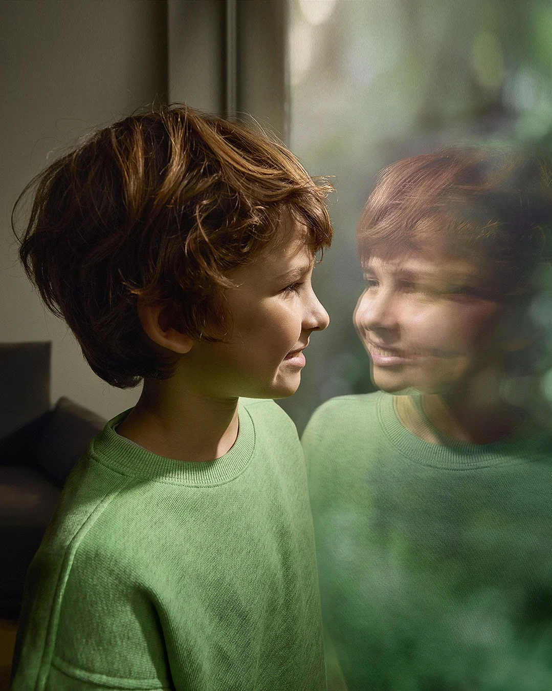 Ein Mädchen mit kurzen braunen Haaren steht vor einem Fenster, schaut sich selbst im Glas an und lächelt.