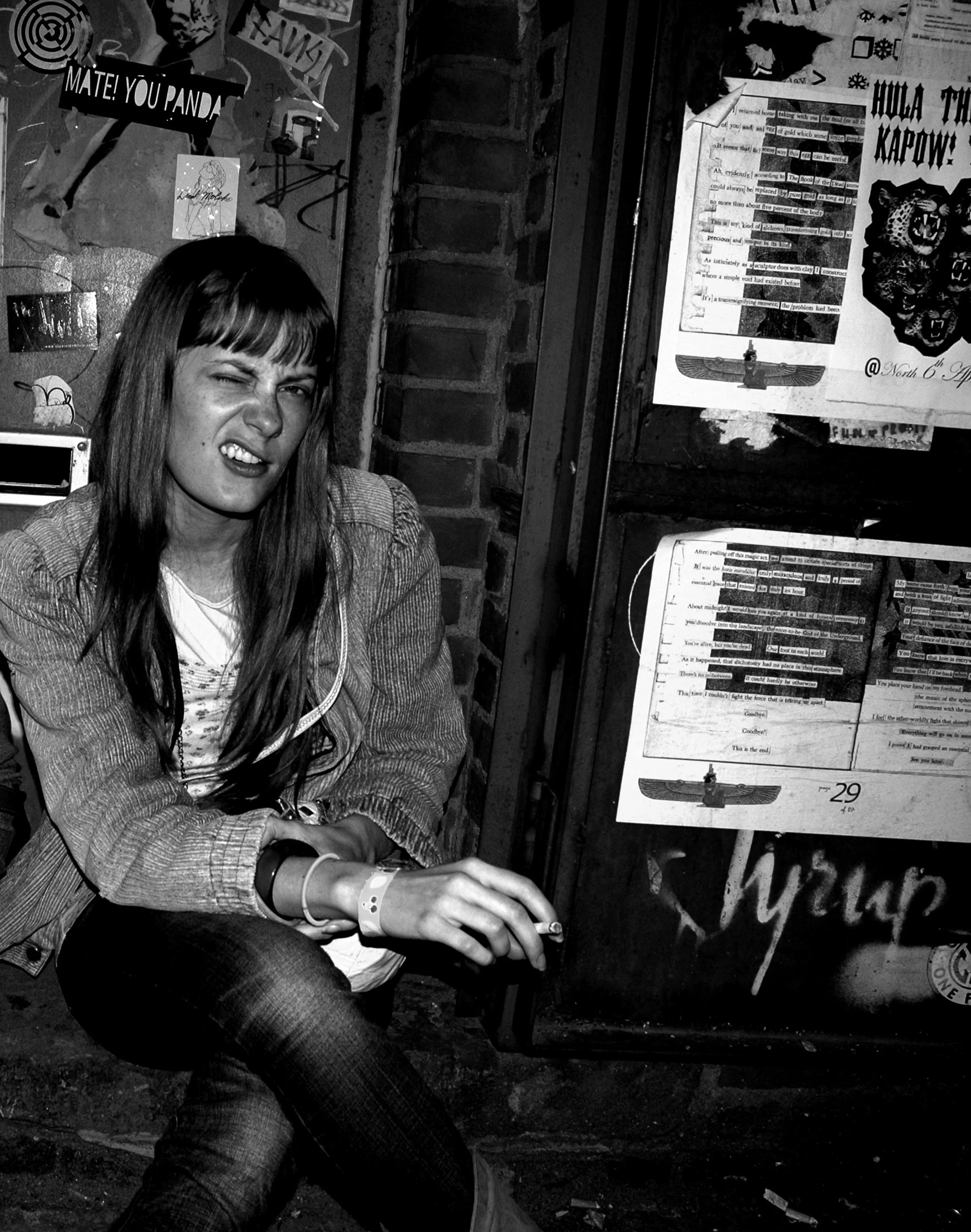 Eine junge Frau macht ein witziges Gesicht, während sie vor einem schwarzen Brett sitzt, das mit Postern und Fotos bedeckt ist.