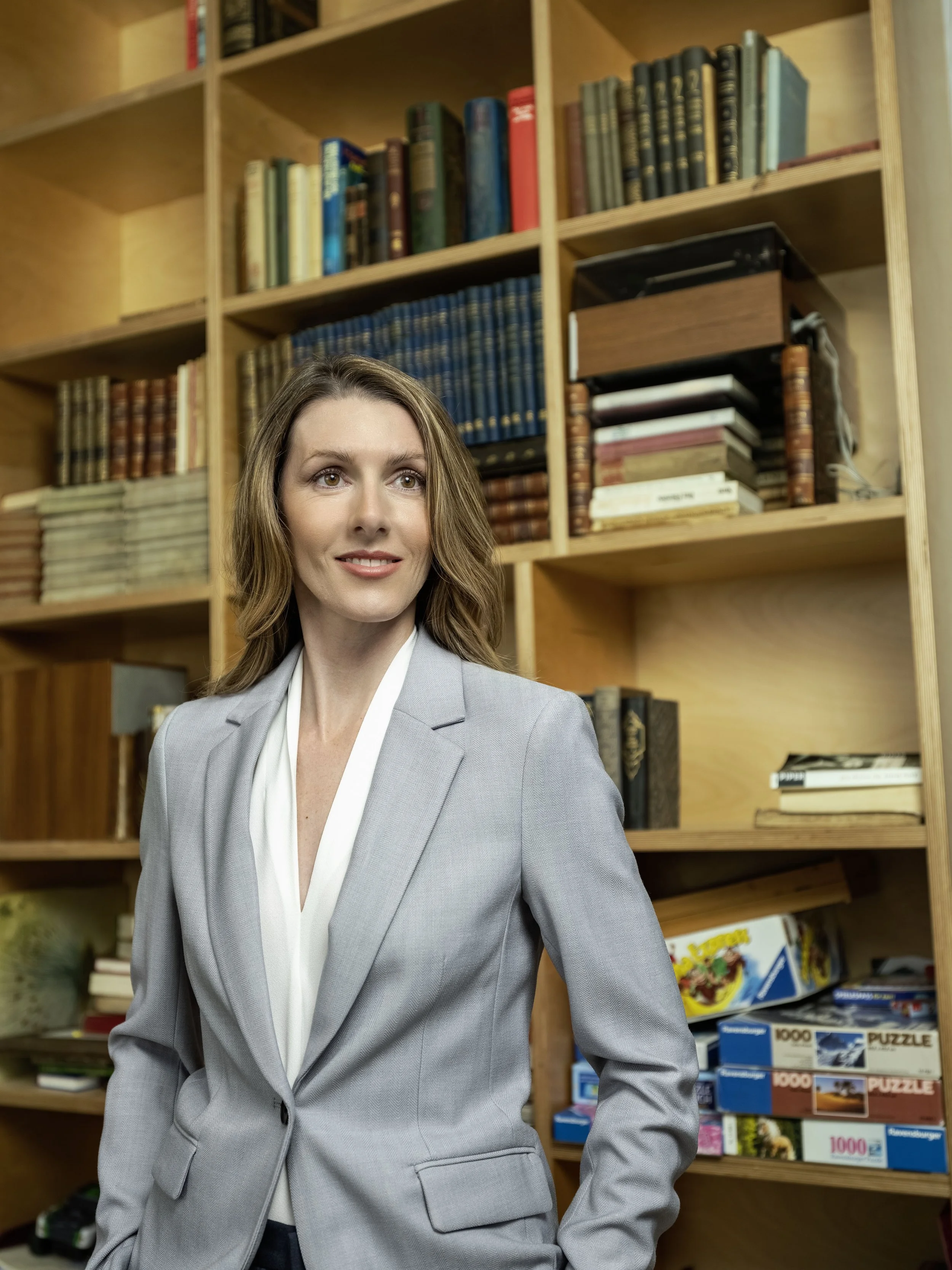 Eine Frau in einem grauen Anzug posiert vor einer Bücherregalwand, die mit Büchern und Puzzles gefüllt ist.