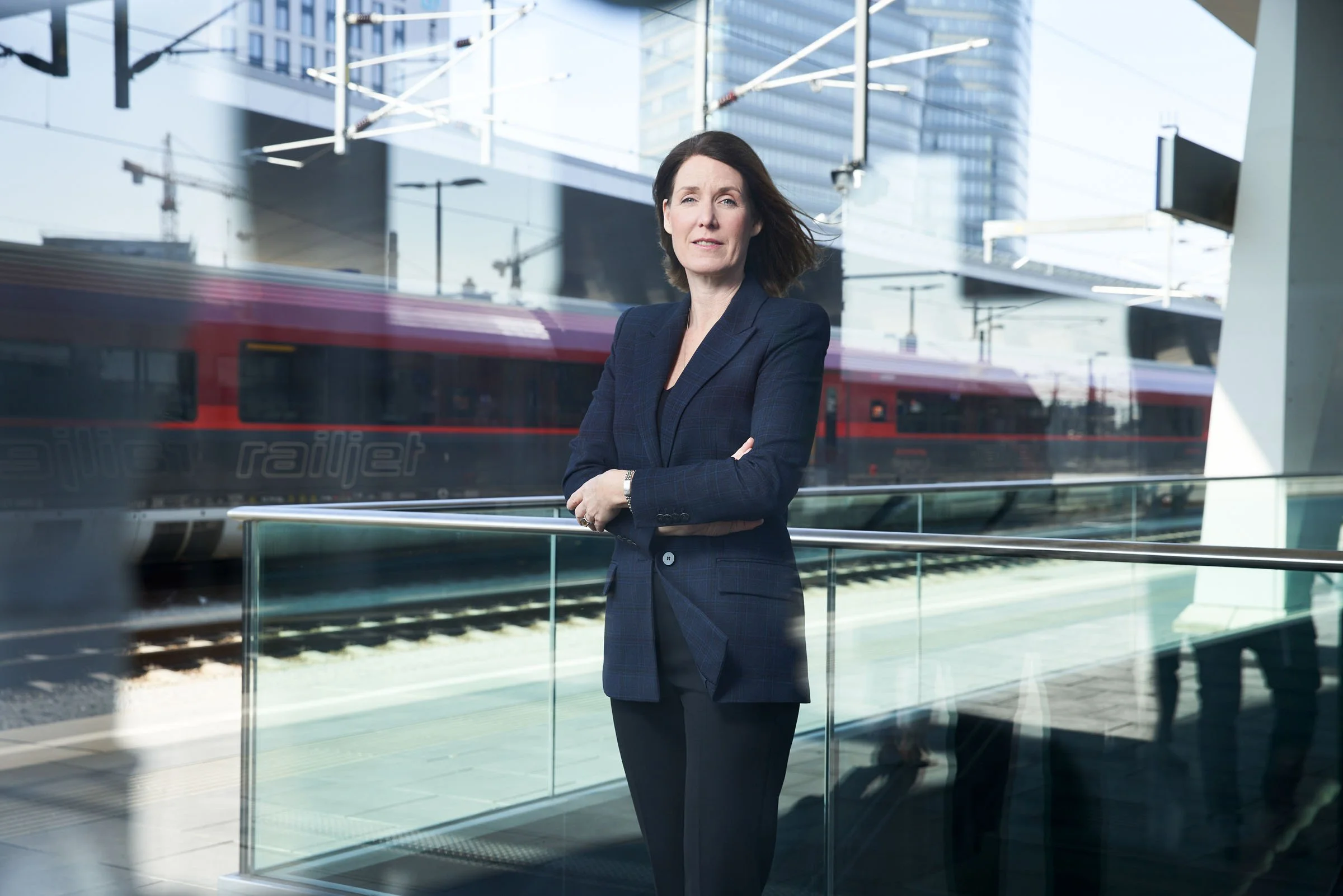 Eine Frau in dunklem Anzug steht an einem Bahnhof mit Bahnlinie im Hintergrund.